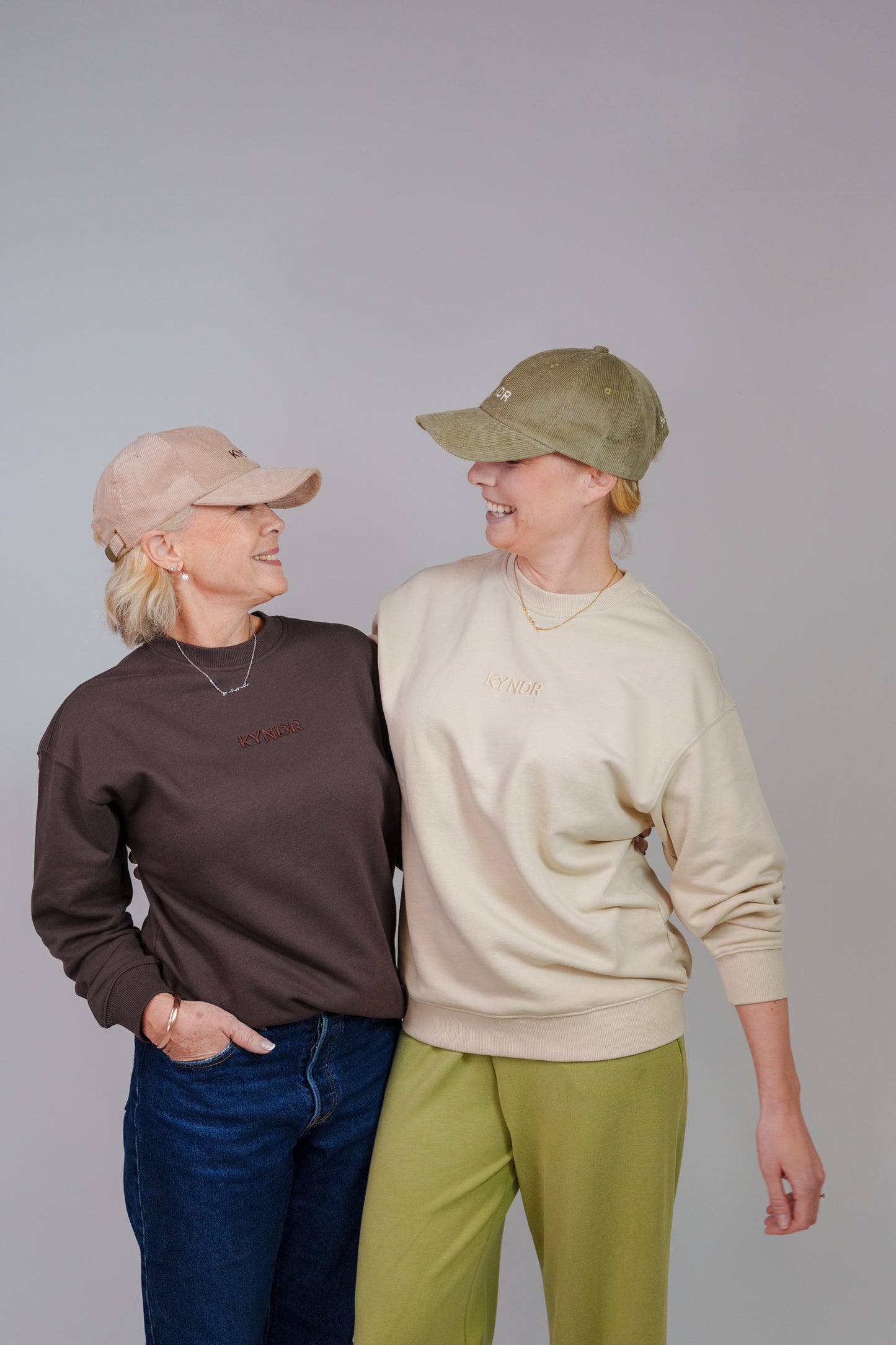 Two women standing and smiling at each other against a plain light gray studio background. The woman on the left wears a chocolate coloured sweatshirt, blue jeans, and a dusty pink cap.
