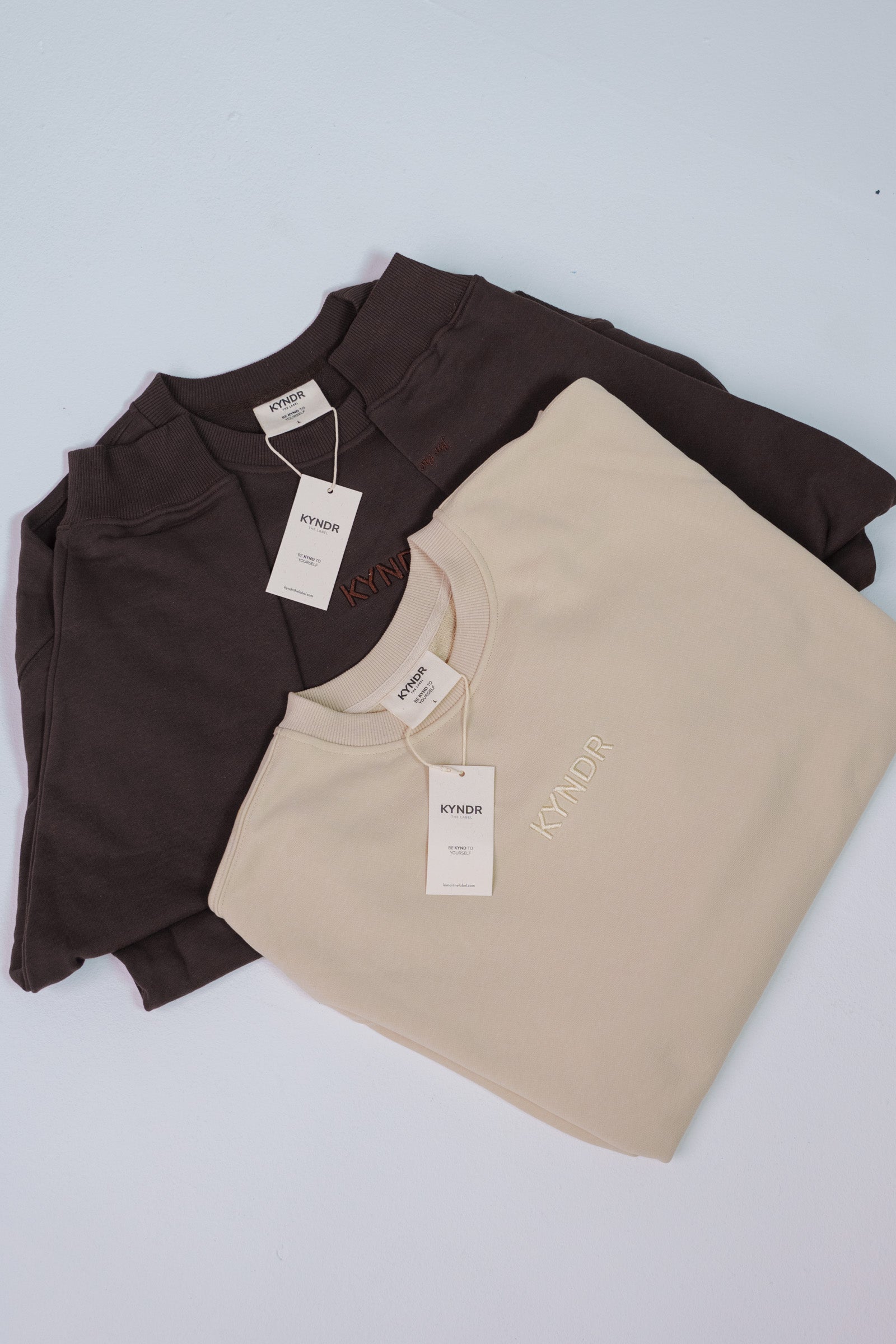 Two folded crewneck sweatshirts from the brand KYNDR, one in chocolate and one in ecru, arranged on a light gray background.
