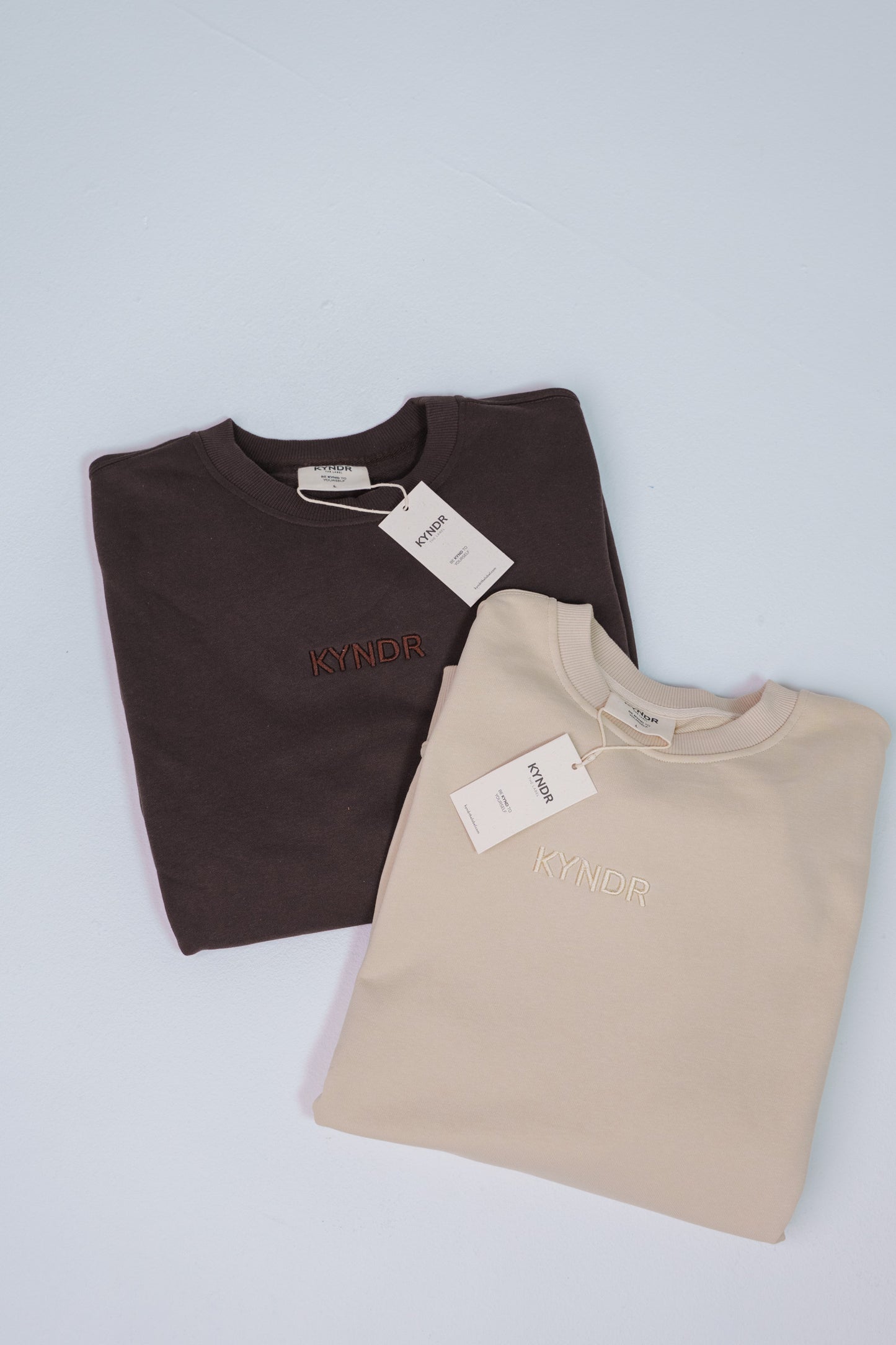 Two folded crewneck sweatshirts on a light gray background, one chocolate with red embroidered text and one ecru with white embroidered text. Both have white tags attached.